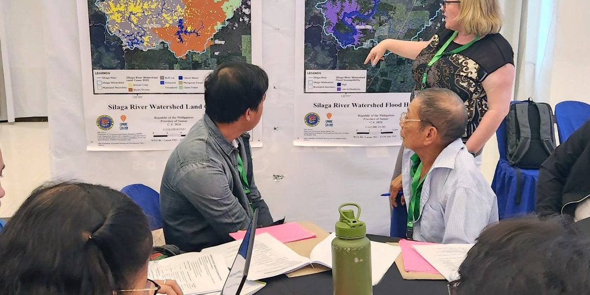 Workshop attendees seated at a table reviewing documents while a facilitator points to large watershed maps displayed on the wall during a collaborative planning session