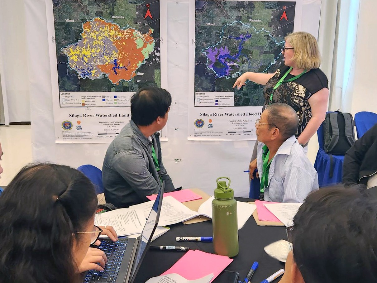 Workshop attendees seated at a table reviewing documents while a facilitator points to large watershed maps displayed on the wall during a collaborative planning session