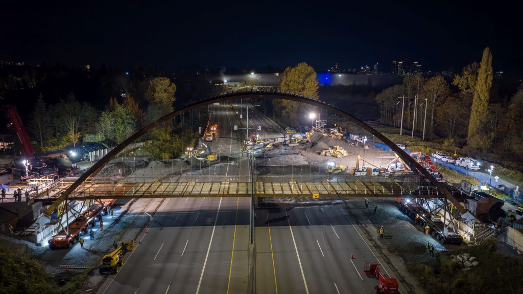 Nighttime construction of the Burnaby Lake Pedestrian Bridge spanning Highway 1, demonstrating bridge erection, construction staging, and transportation infrastructure delivery.