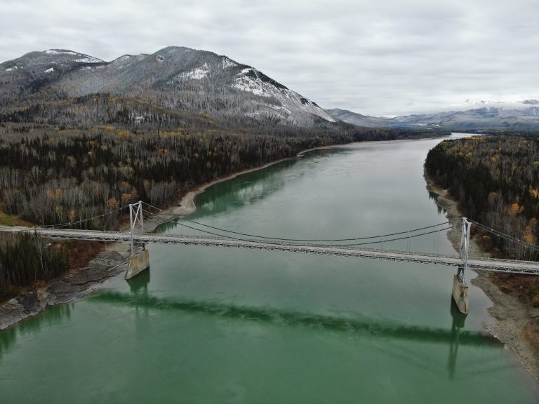 Aerial view of a suspension bridge crossing a large river surrounded by mountainous terrain and boreal forest along the Alaska Highway in British Columbia.