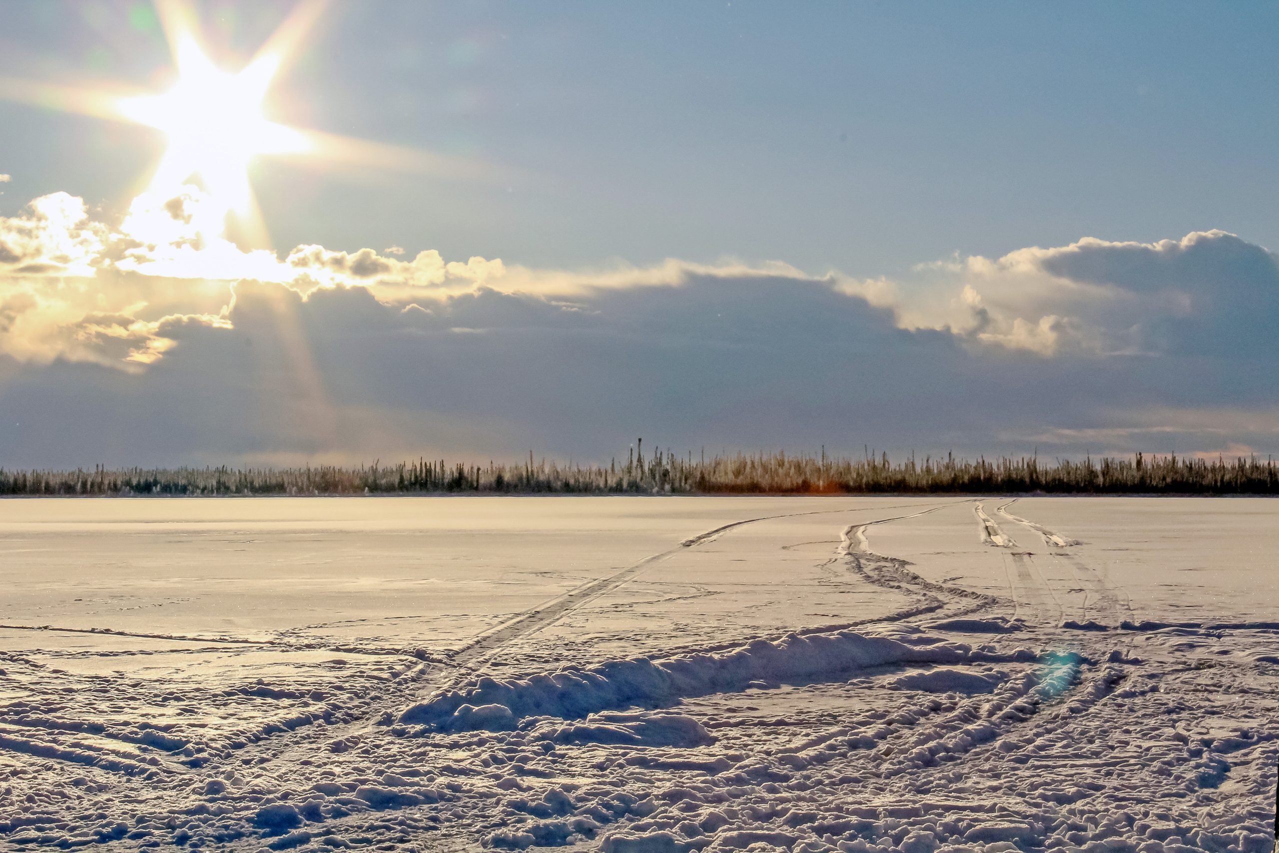 Snow-covered landscape near Fort Nelson with vehicle tracks leading toward a treeline under a bright winter sun