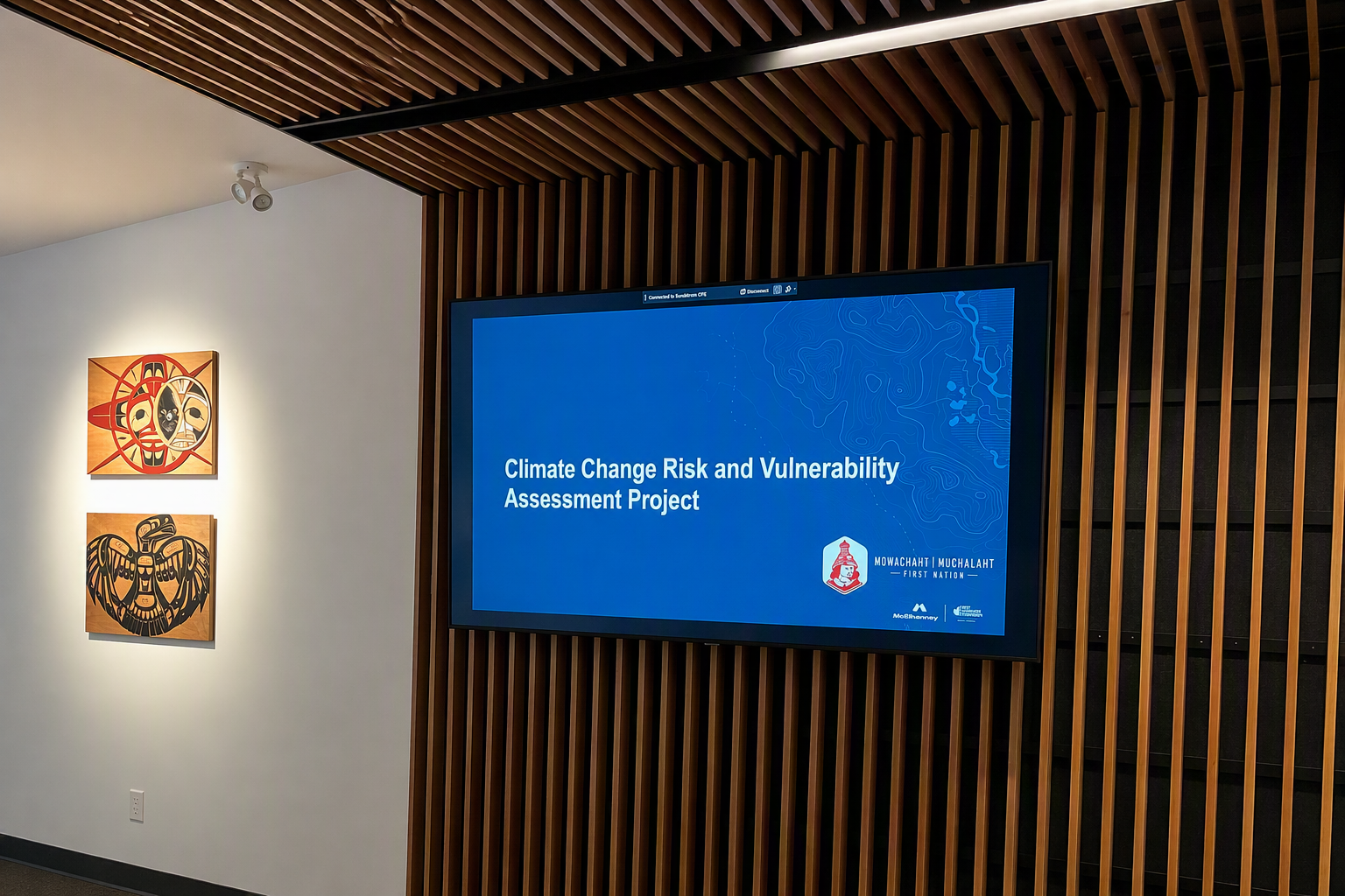 Mowachaht Muchalaht First Nation Climate Change Risk and Vulnerability Assessment workshop Wall-mounted display showing a Climate Change Risk and Vulnerability Assessment presentation for Mowachaht Muchalaht First Nation inside a meeting space with Indigenous artwork and wood architectural features.